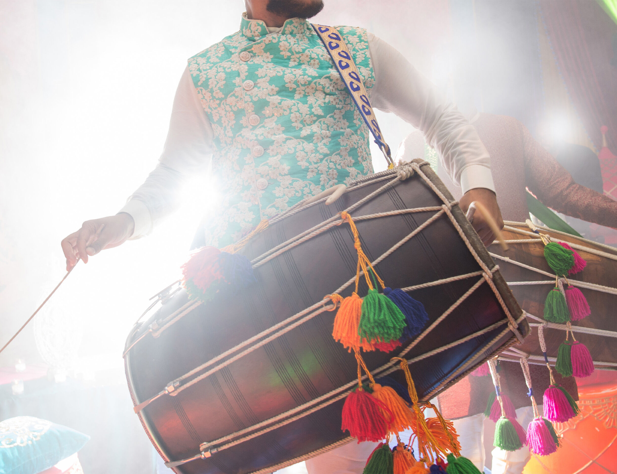Punjabi man playing a traditional dhol drum with colorful tassels during Baisakhi celebration, wearing festive ethnic attire and surrounded by bright lights.